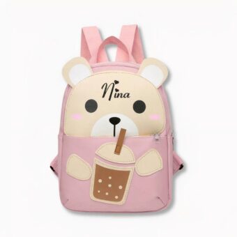 Kleine roze rugzak voor kinderen in de vorm van een teddybeer die nipt van een glas met een rietje waarop de naam Nina staat, op een lichtgrijze achtergrond
